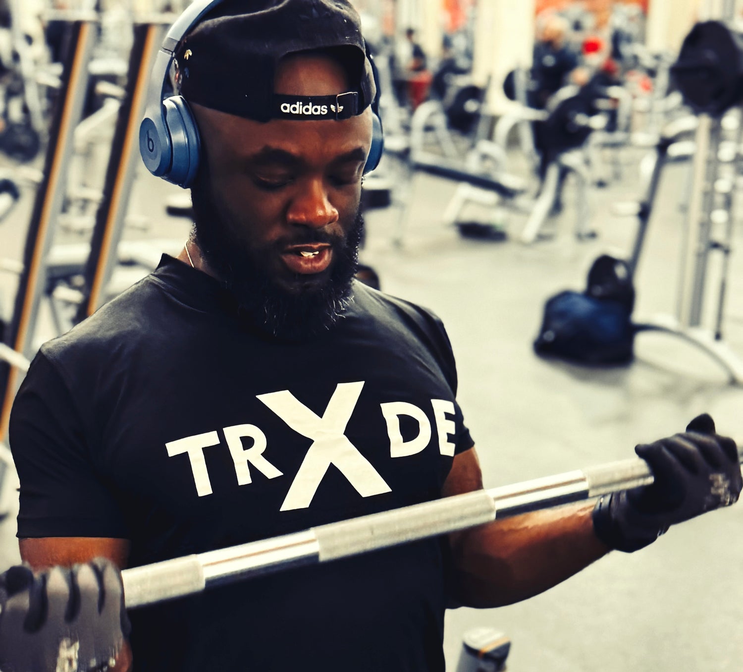 Person wearing a black 'TRX DE' shirt and headphones, holding a barbell in a gym setting.