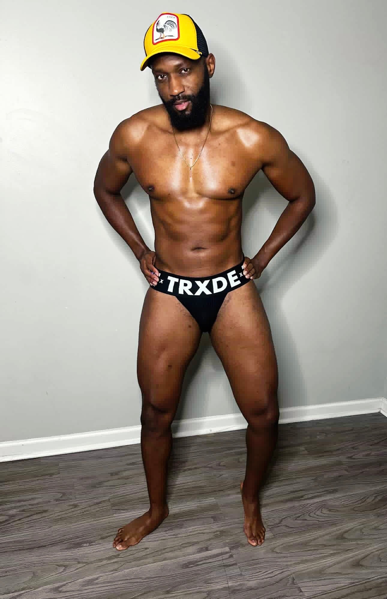 Man wearing a yellow cap and black underwear with 'TRXDE' branding against a plain wall.
