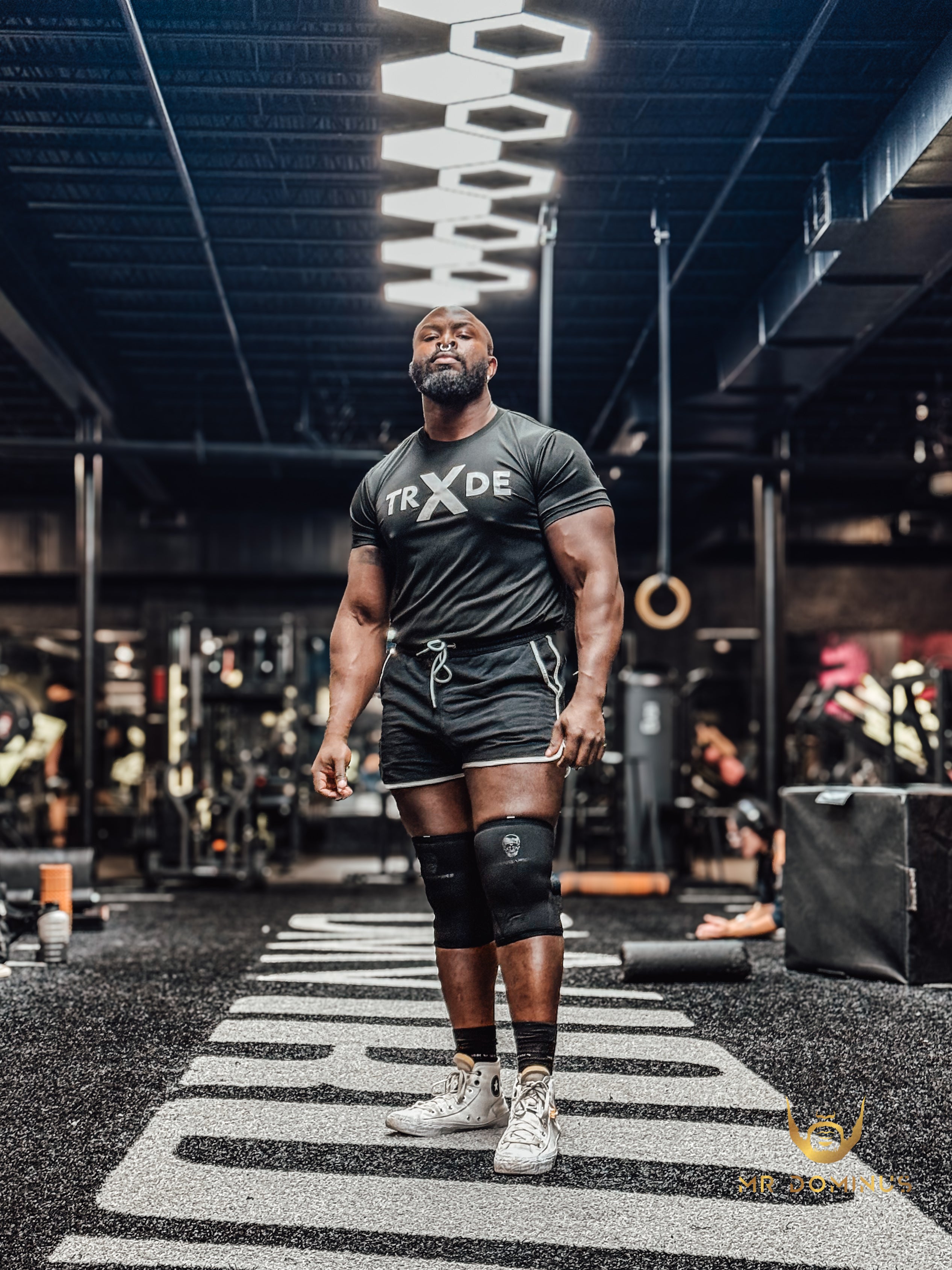 Man in a gym wearing a TRX shirt and shorts, standing on a striped floor.