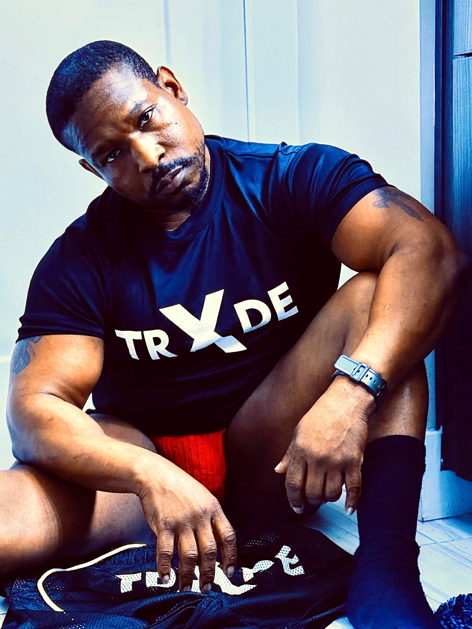 Black man wearing a black shirt with white text, red underwear, and sitting on black shorts