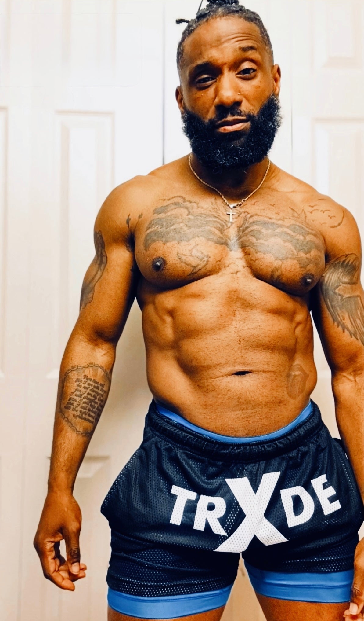 A muscular man with a beard and tattoos stands shirtless wearing dark navy athletic shorts with blue trim and a large white 'TRXDE' logo on the front, photographed indoors against a light door background.
