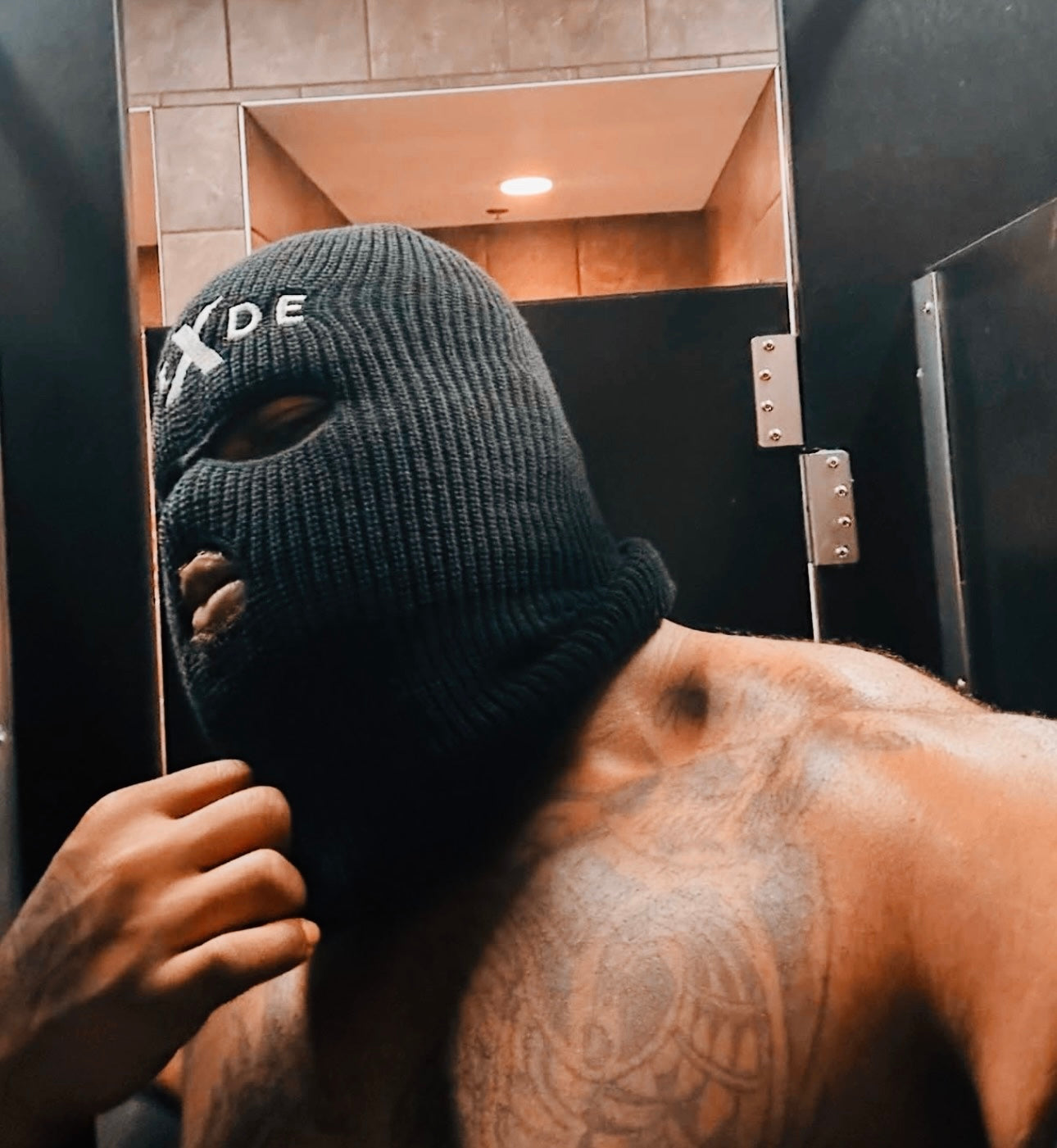 Person wearing a black ski mask with a logo, standing in a bathroom.