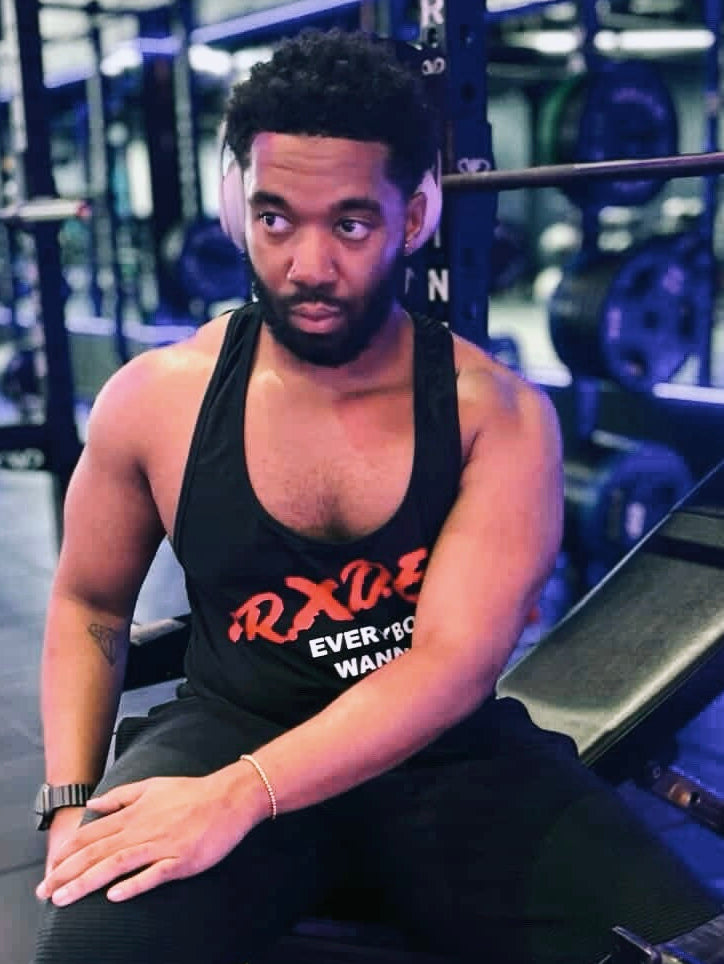 Man sitting on a weight bench in a gym wearing a black tank top with orange text.