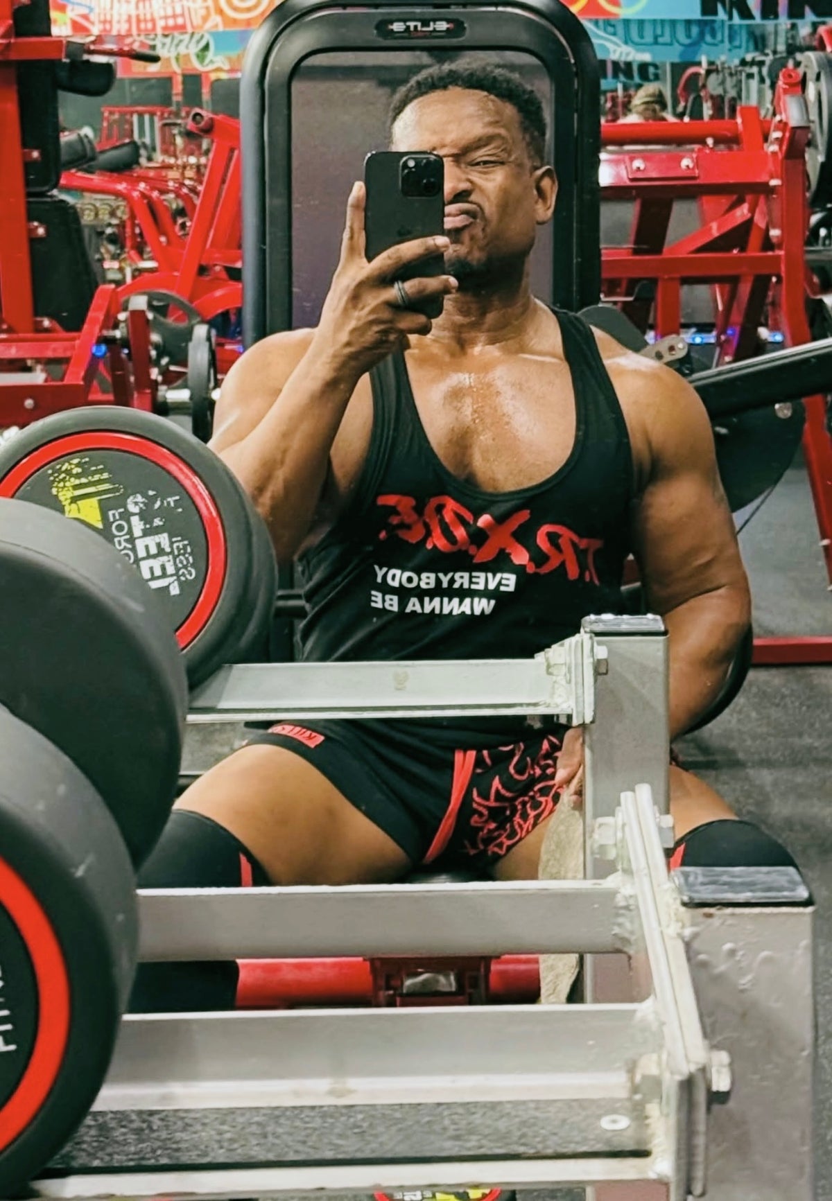 Man taking a mirror selfie in a gym wearing a black tank top with red text.