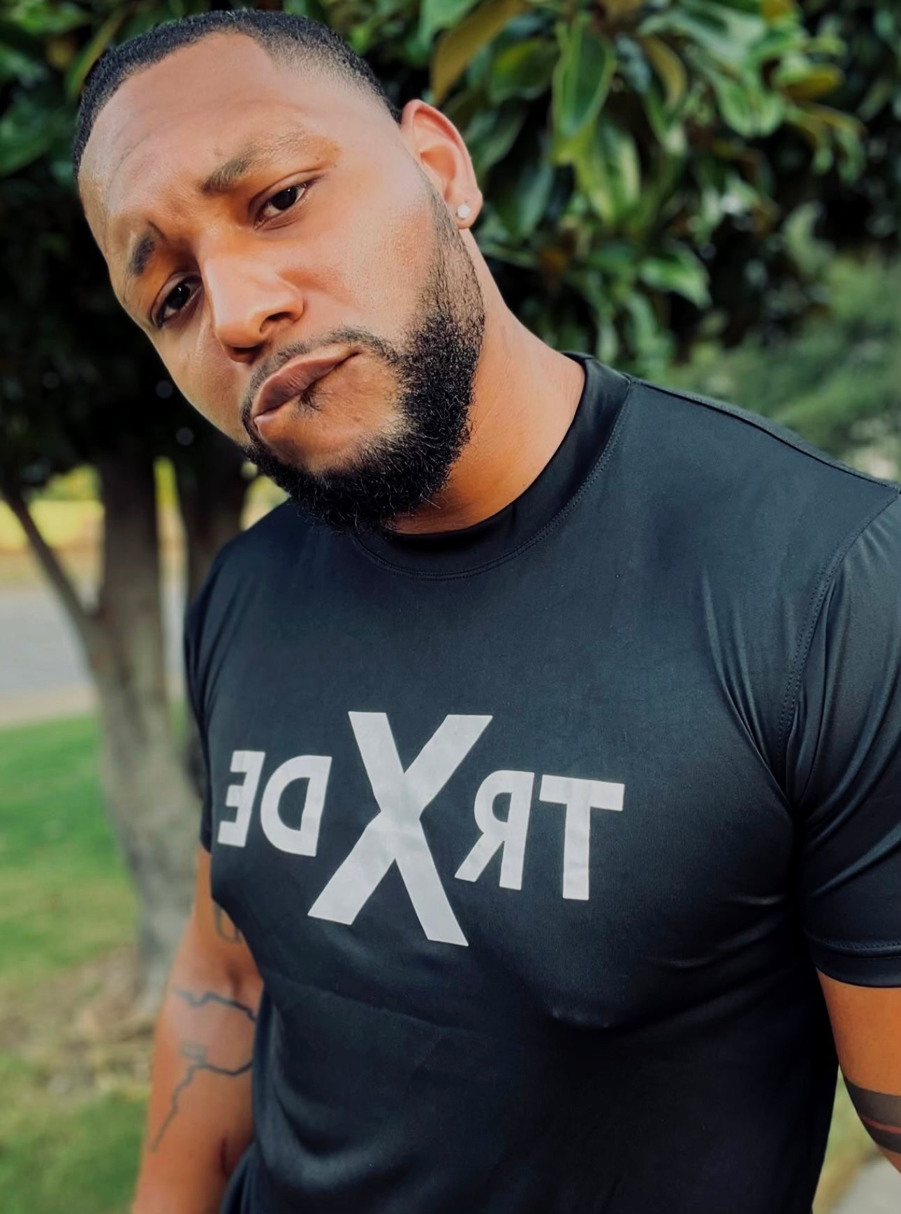 A man wearing a black athletic t-shirt with a large white mirrored 'TRX' logo on the chest, photographed outdoors with trees in the background.