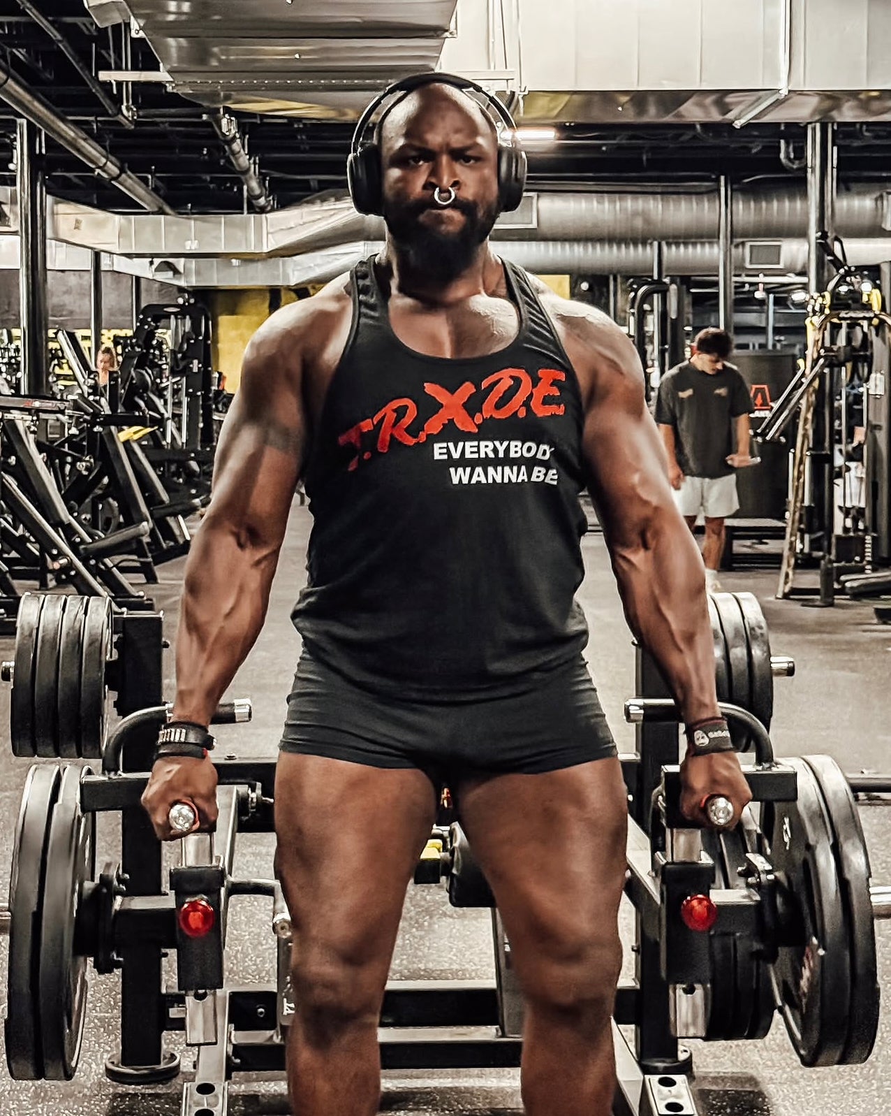 A muscular man in a gym wearing a black tank top with red and white text that reads 'TRXDE EVERYBODY WANNABE' and black shorts, standing in front of a barbell machine.