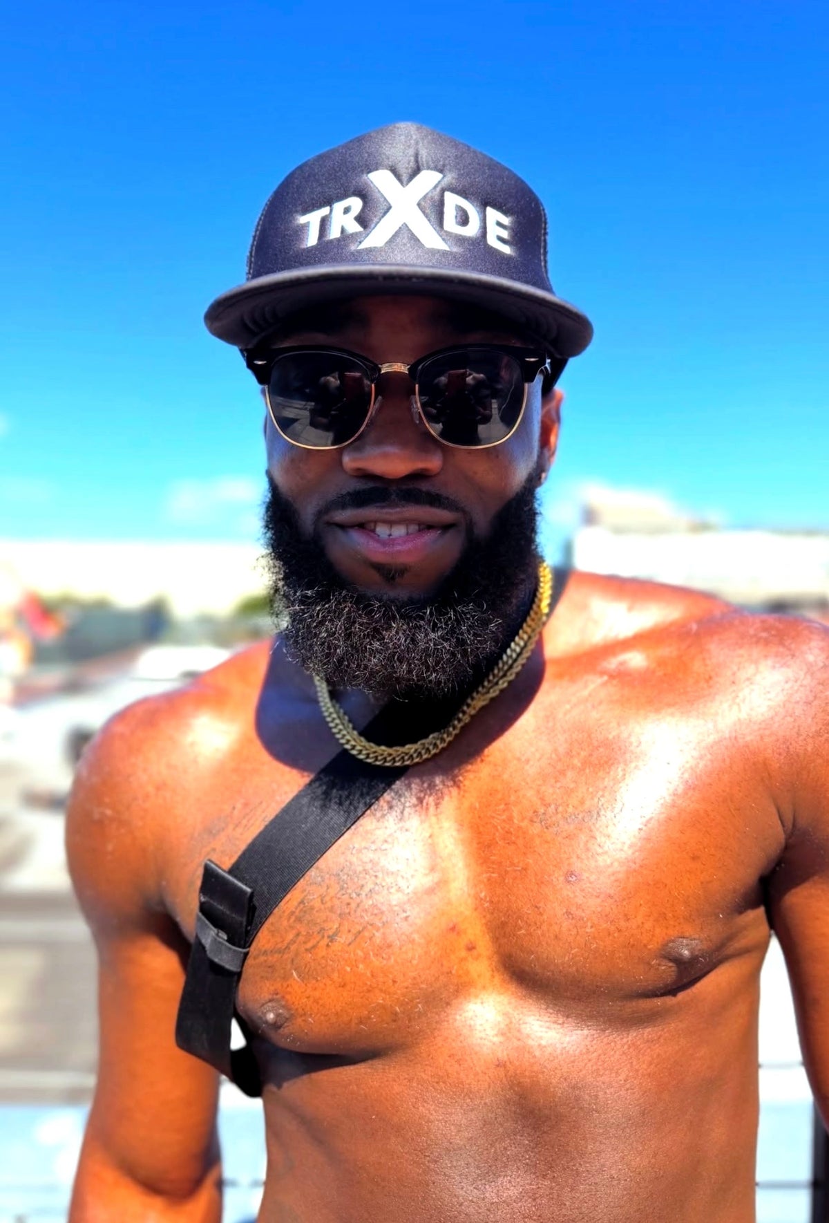 A shirtless man wearing a black baseball cap with 'TRXDE' in white, gold-framed sunglasses, and a gold chain necklace, photographed outdoors against a bright blue sky.